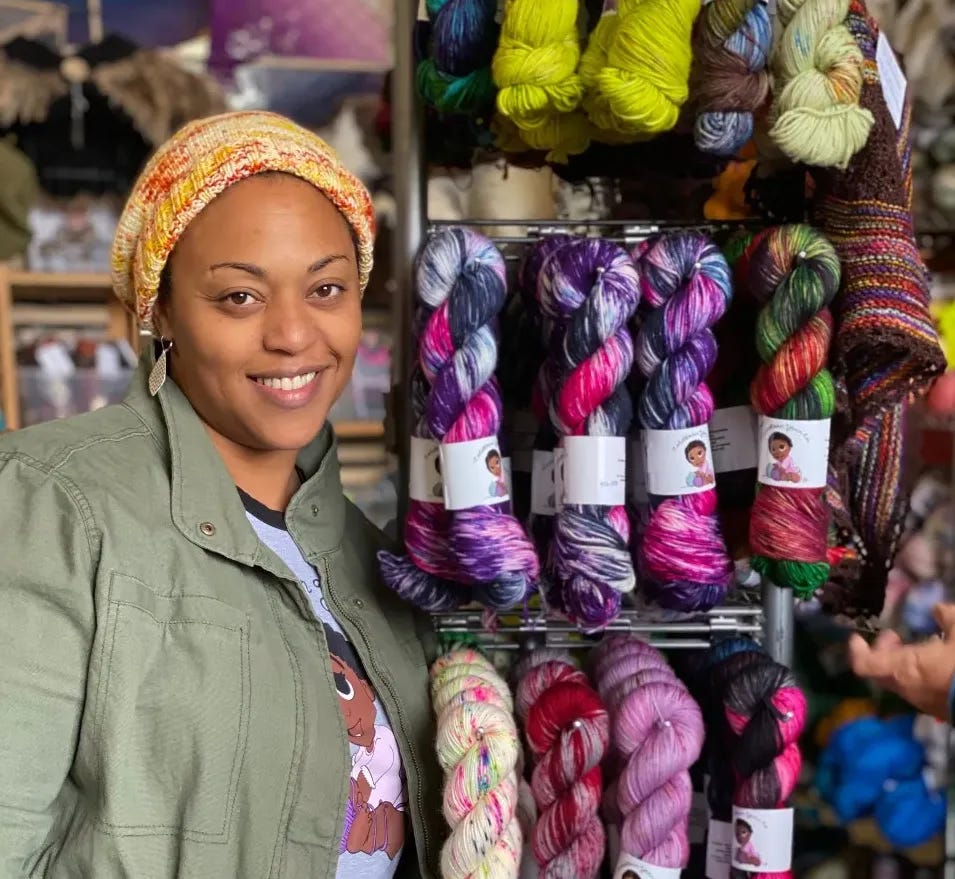 a smiling woman wearing a knitted hat and an olive green jacket stands next to a display rack of brightly colored yard skeins 