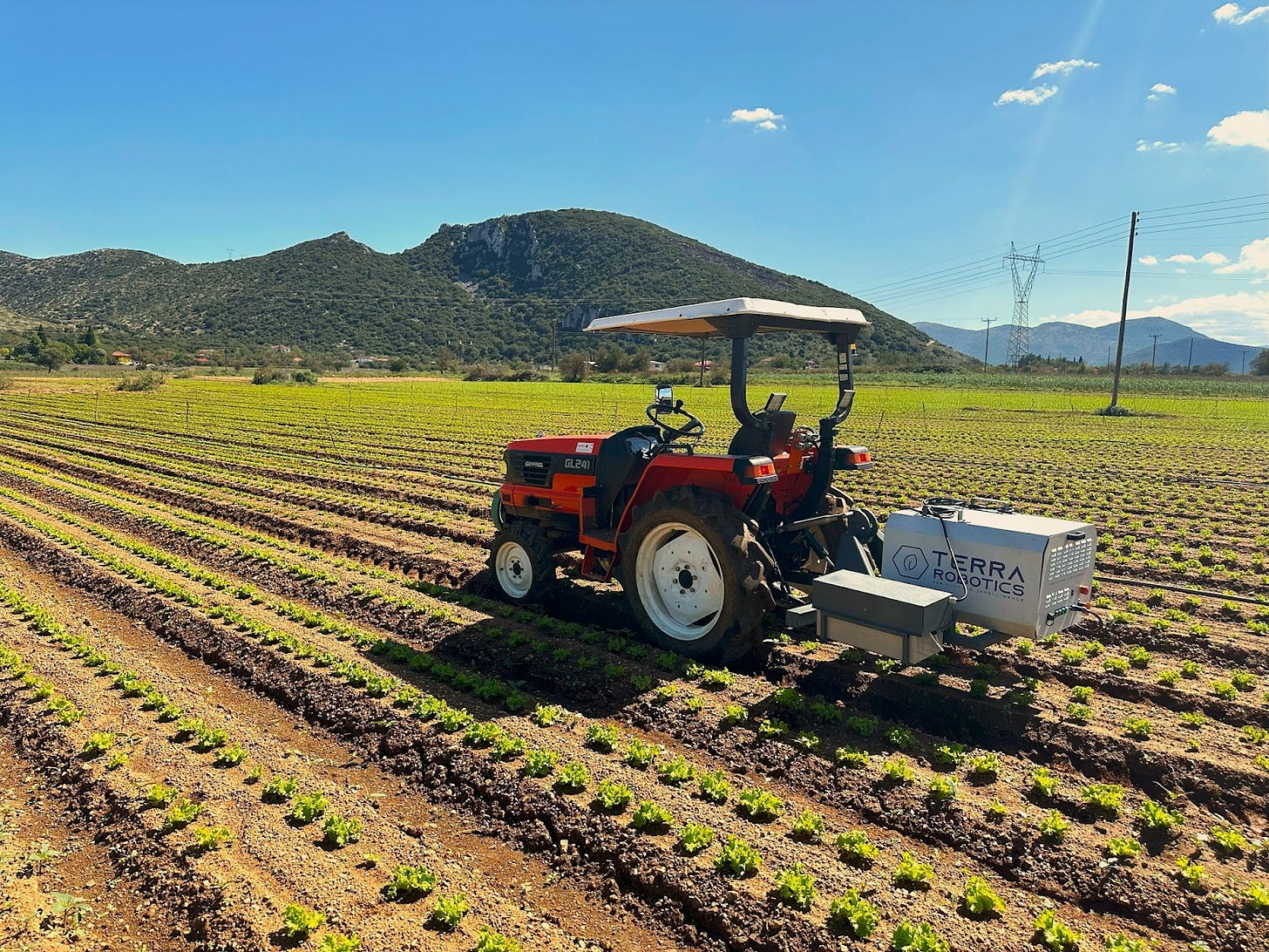 Tractor with a Terra Robotics unit attached operates in a sunny vegetable field with neat crop rows, surrounded by mountains and blue sky in the background