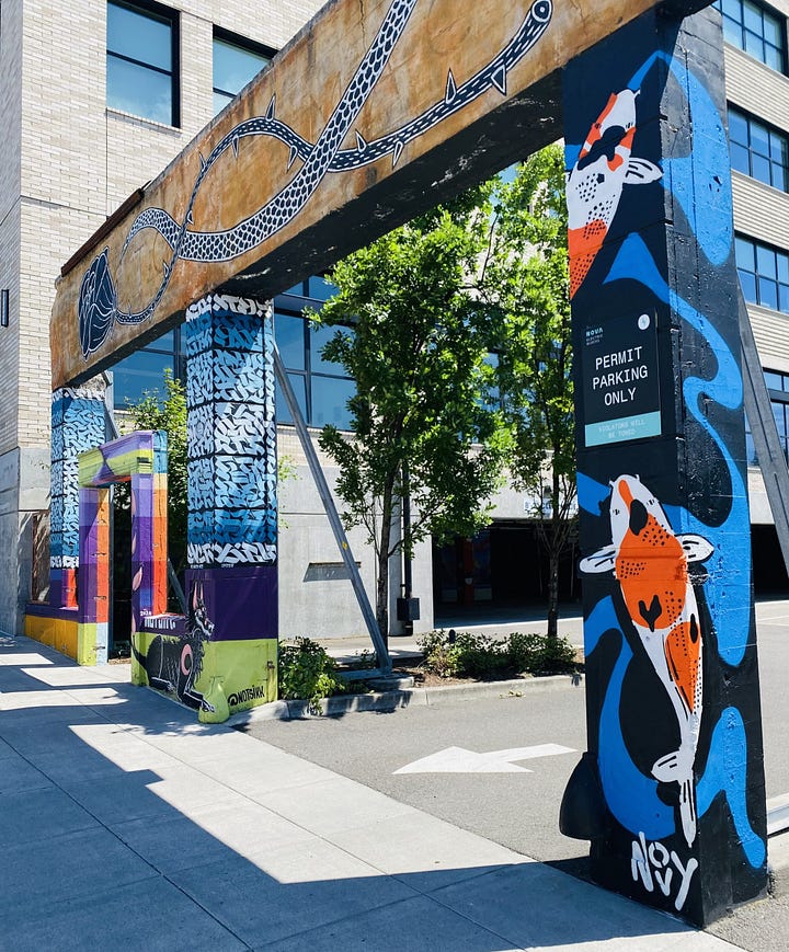 1) Jeremy is wearing a colorful shirt and bandana standing in front of a vibrant graffiti mural of a red brick with "PRIDE!" in rainbow letters, on top of a black wall, evoking a sense of celebration and diversity. Jeremy's signature and tag, Novy, is in white in the bottom right corner. Jeremy is wearing a white T-shirt with a clown face on it, and blue jeans. He is wearing a blue baseball cap and a black bandana over his nose and mouth. He has his thumbs in his pants pockets. 2) Colorful urban street art on a concrete structure, featuring a blue curled pattern, koi fish, and abstract designs. Nearby trees and parking sign visible. Jeremy's koi are on the far right pillar, and his signature and tag are on the bottom right of the pillar. It is attached to a white. building with several windows.