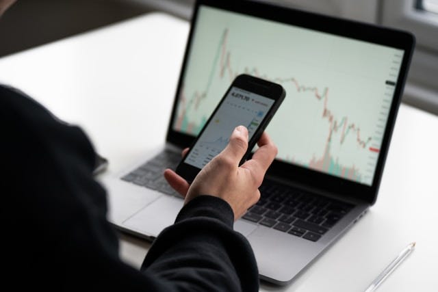 A computer, a phone, an investment portfolio with the investor looking at both A computer, a phone, an investment portfolio with the investor looking at both