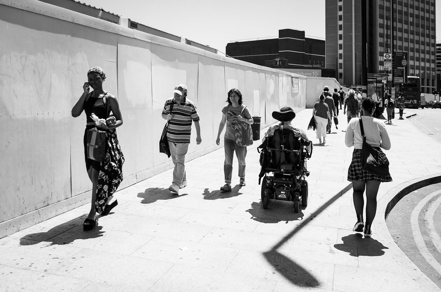 People walk along a sun-lit road alone or in small groups. One is Jamie Hale, wheeling away from the camera in their electric wheelchair