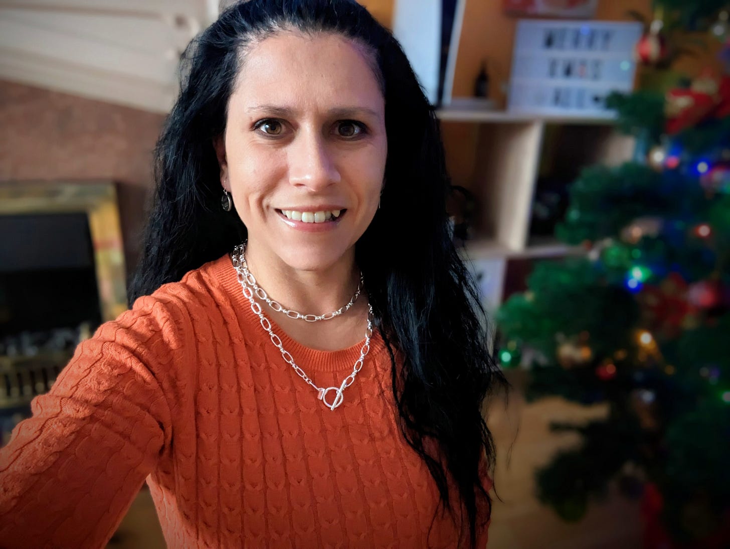 Teo Byrne of SENDwise Hub. She is a lady with black hair, wearing an orange jumper and a silver necklace. There is a Christmas tree and a fireplace in the background.