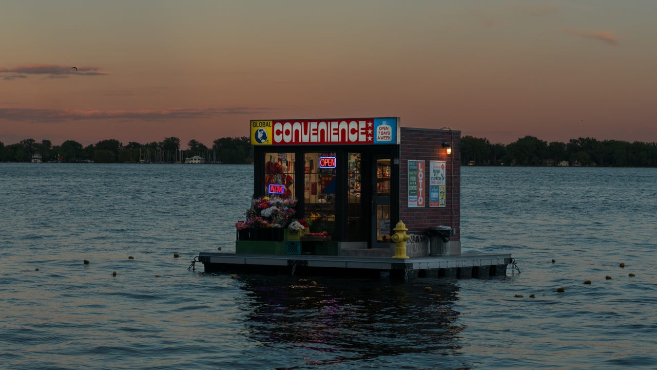 A small convenience store floats on the water at sunset, its neon “Convenience” and “Open” signs glowing, with flower displays outside and a calm shoreline in the background. A small convenience store floats on the water at sunset, its neon “Convenience” and “Open” signs glowing, with flower displays outside and a calm shoreline in the background.