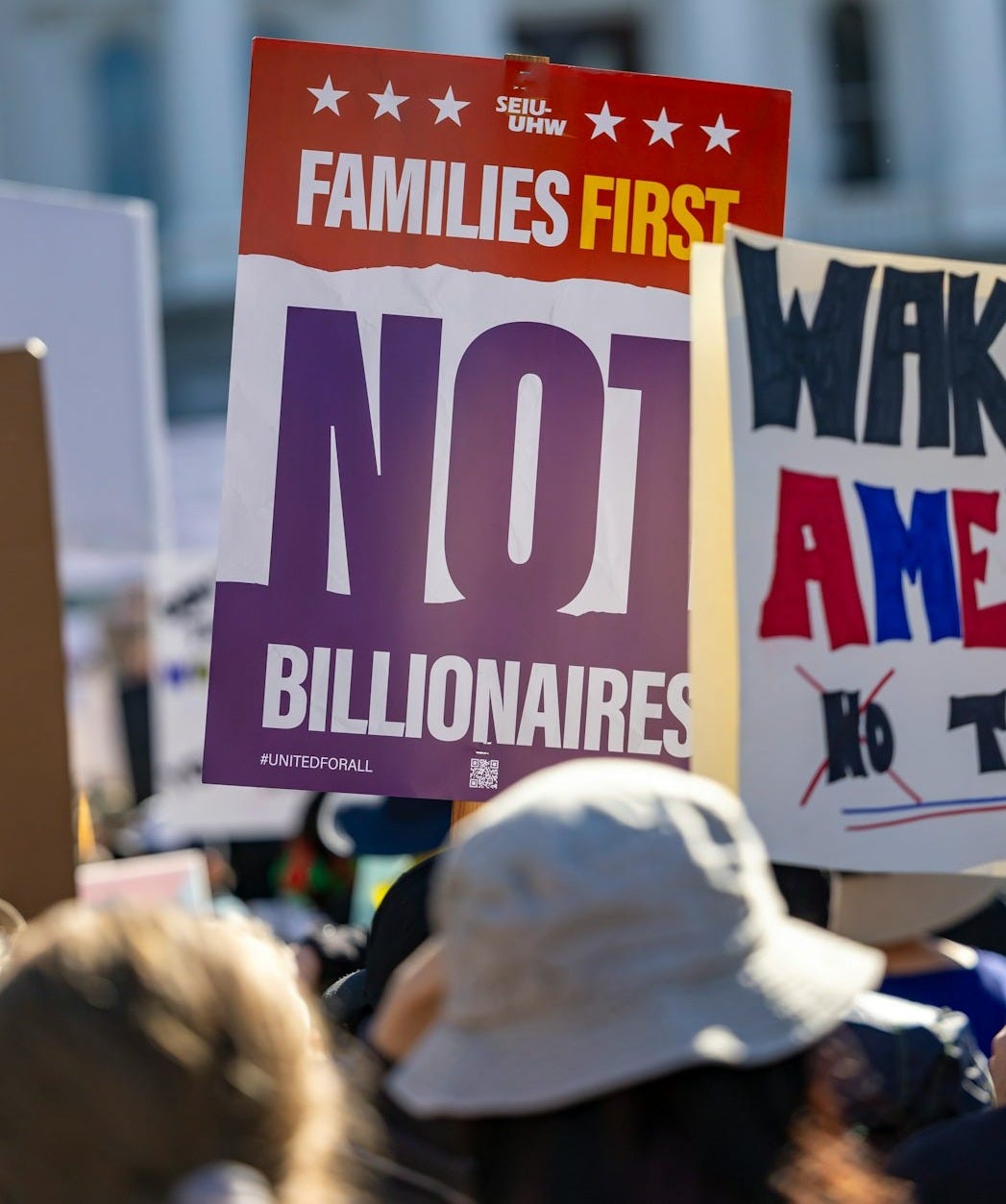 Protest signs reading 'families first, not billionaires' and 'wake america'