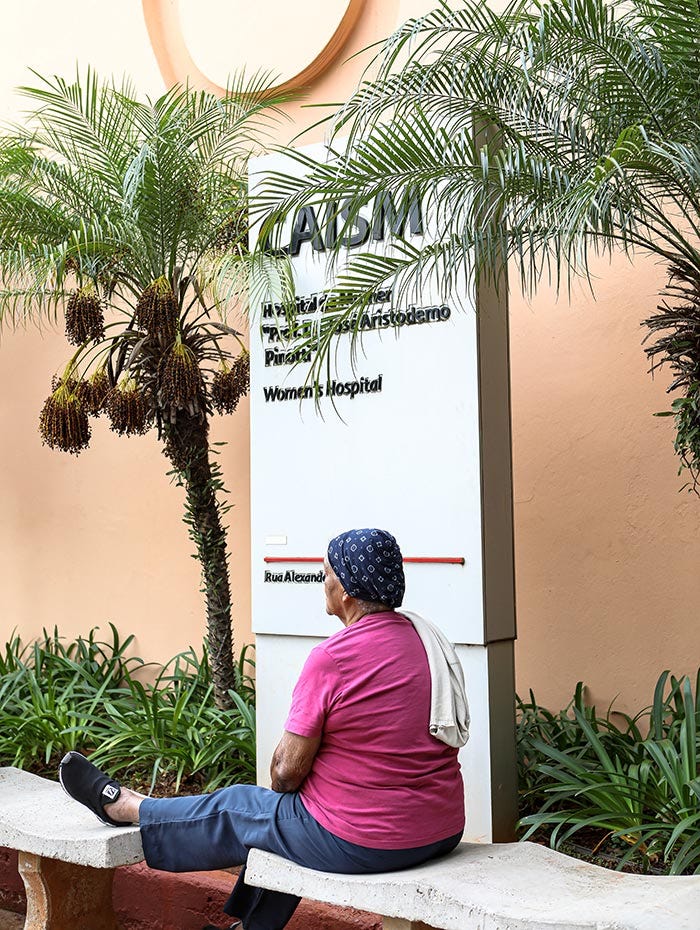 Pessoa sentada em banco de concreto, cercada por vegetação tropical incluindo palmeiras, em ambiente externo com parede em tons de pêssego ao fundo.