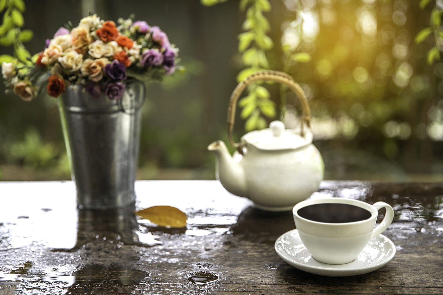 A white tea cup of and a saucer, a white teapot and a metal bucket of colorful flowers, sit on a wet wooden table, just after the rain. There’s greenery and sunlight in the background.