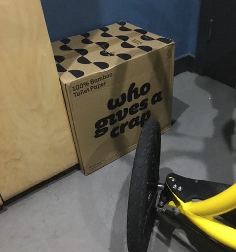 Interior of an accessible toilet, also being used to store boxed toilet paper from the 'who gives a crap' brand. Jane's yellow Alinker walking bike is also partially in view. Interior of an accessible toilet, also being used to store boxed toilet paper from the 'who gives a crap' brand. Jane's yellow Alinker walking bike is also partially in view.