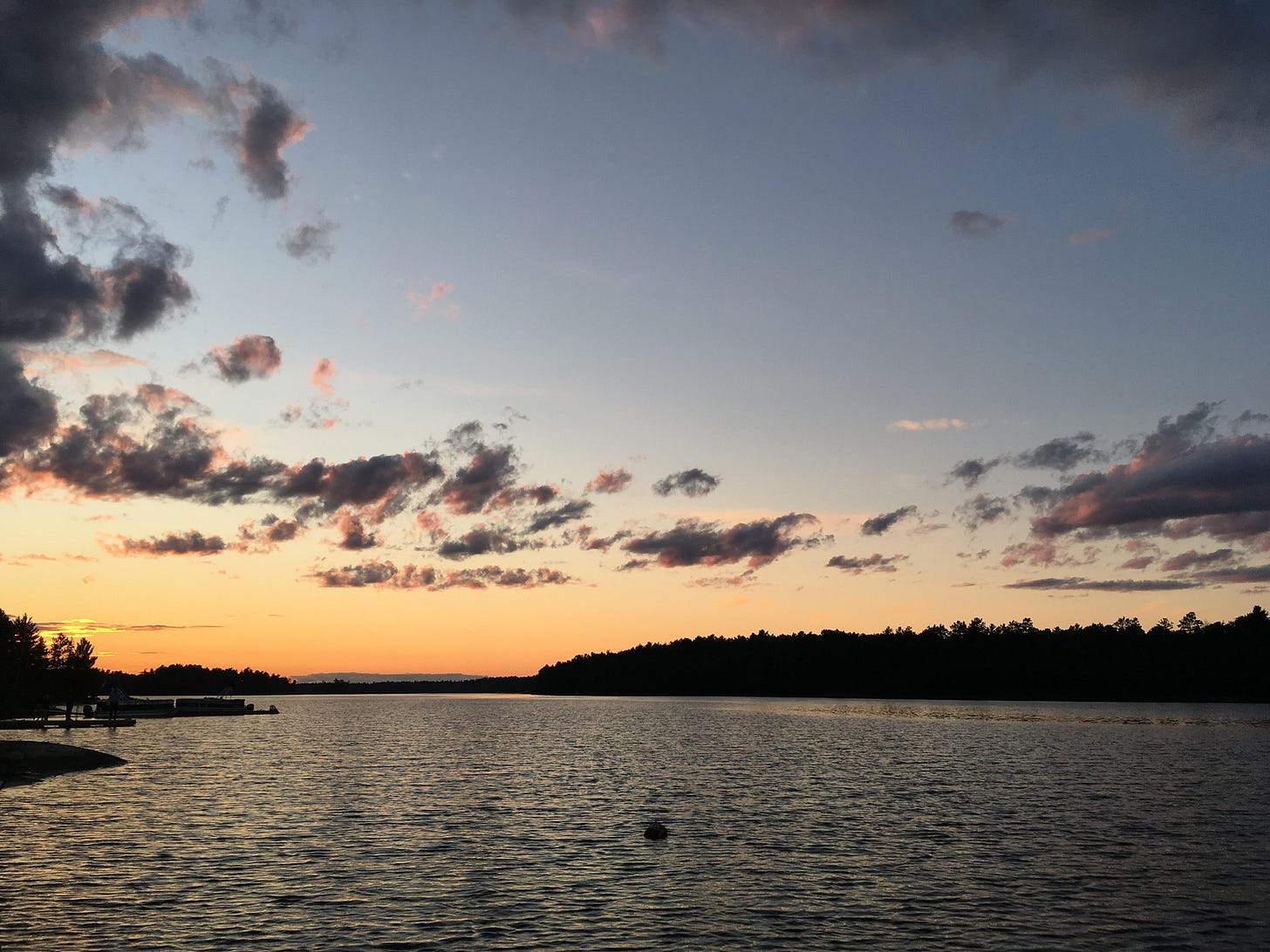 A calm lake at sunset with a tree-lined shoreline and scattered clouds in the deep blue and orange sky. A calm lake at sunset with a tree-lined shoreline and scattered clouds in the deep blue and orange sky.
