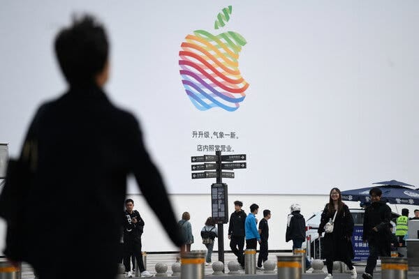 A photograph of a sidewalk with several people walking in different directions, and street signs written in Chinese characters. In the background a large white poster bears the multicolor Apple Inc logo above small black Chinese characters. 