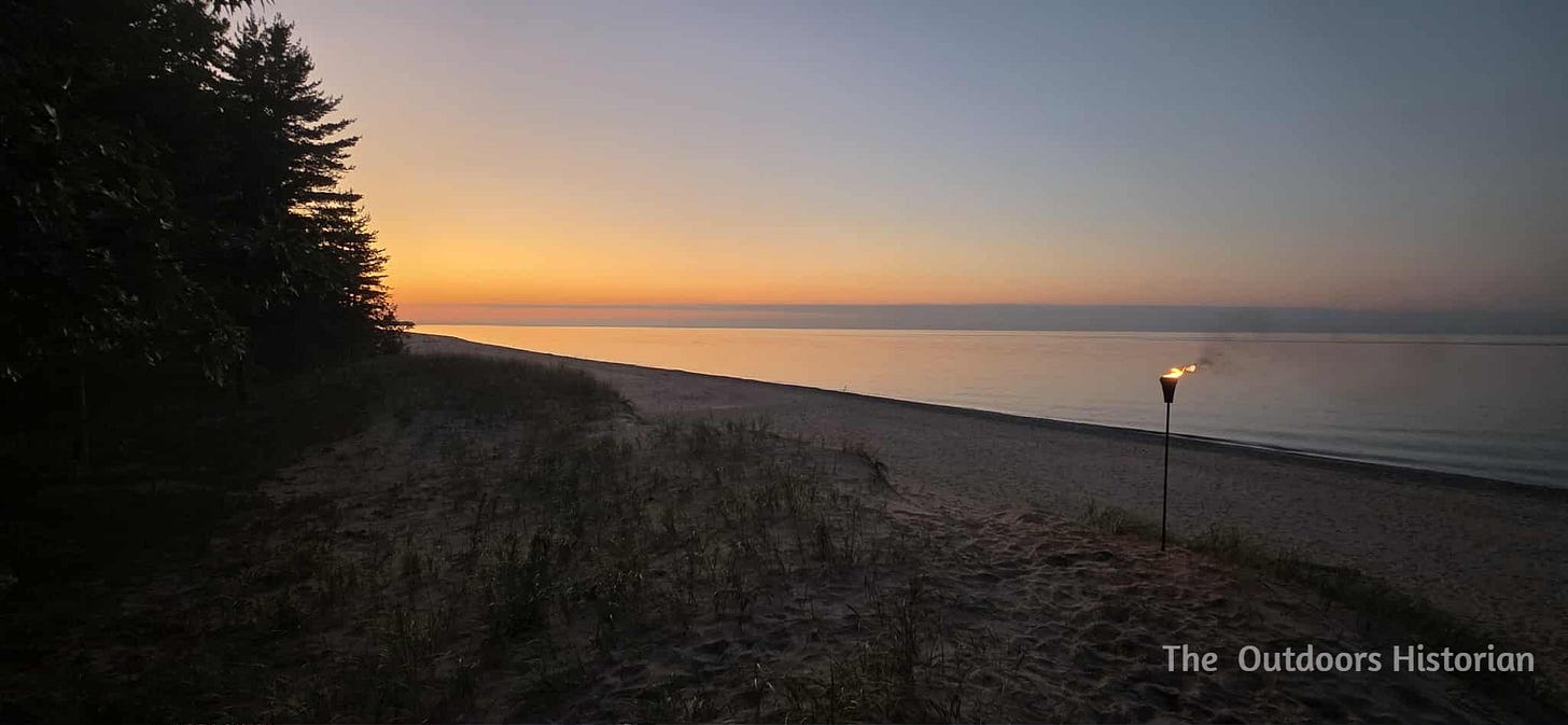 The setting sun creates a warm glow across the sky, reflecting off a calm Lake Superior. The view is from the edge of a low dune with tiki torch near a path down to the beach.