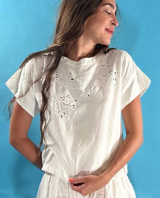 May include: White two-piece outfit featuring a short-sleeved top with floral cutouts and a tiered mini skirt. The outfit is made of a lightweight fabric, perfect for warm weather. The background is a solid light blue.