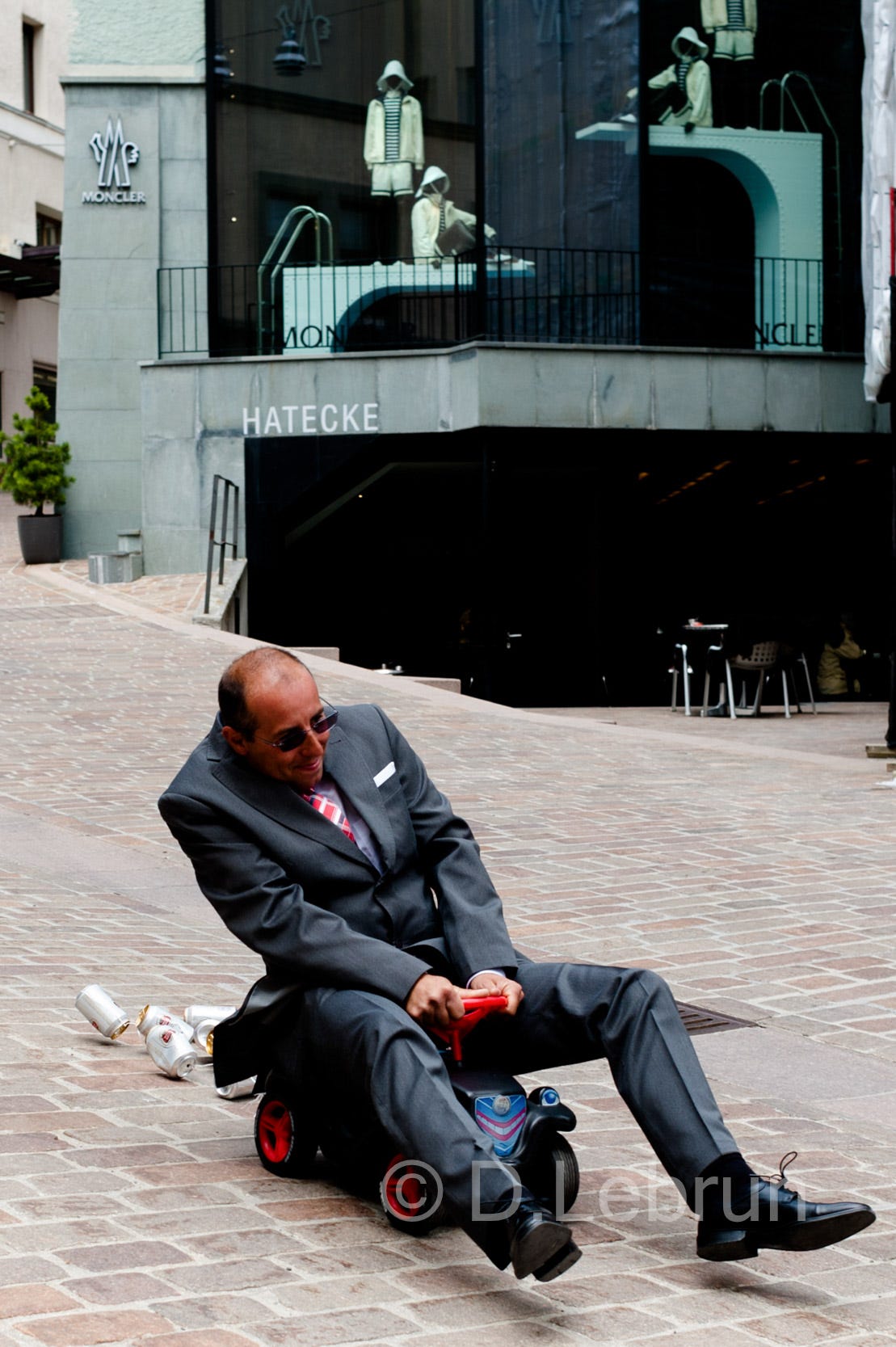 A groom in an expensive looking suit sits on a baby roller with tins attached behind and races down a street in St. Moritz, while in the background faceless women puppets sit in a showcase window, 2013, Switzerland (photo/Dorothée Lebrun). 