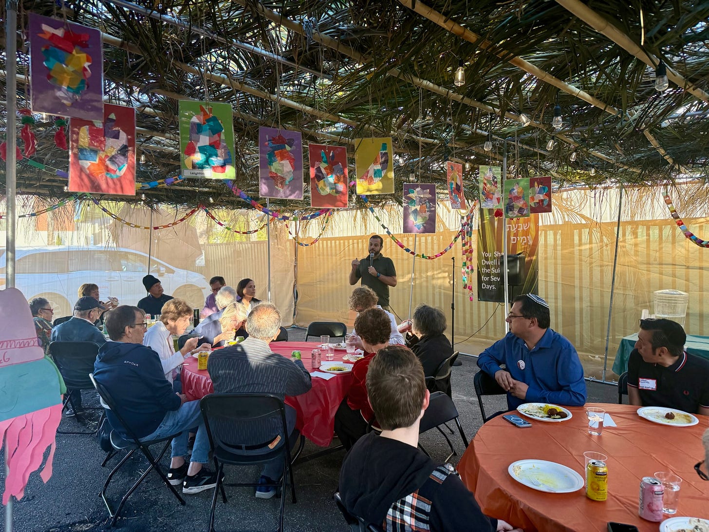 A group of diverse people sit in a sukkah - an outdoor structure with temporary tan fabric walls, a ceiling made of green palm leaves, with paper stained glass pictures along the roof and paper chains lining the walls. The people are sitting at tables covered in red, orange, and green tablecloths, and each person has a plate of food and a drink in front of them. They are all attentively facing a white man with dark brown hair holding a microphone at the front of the structure.