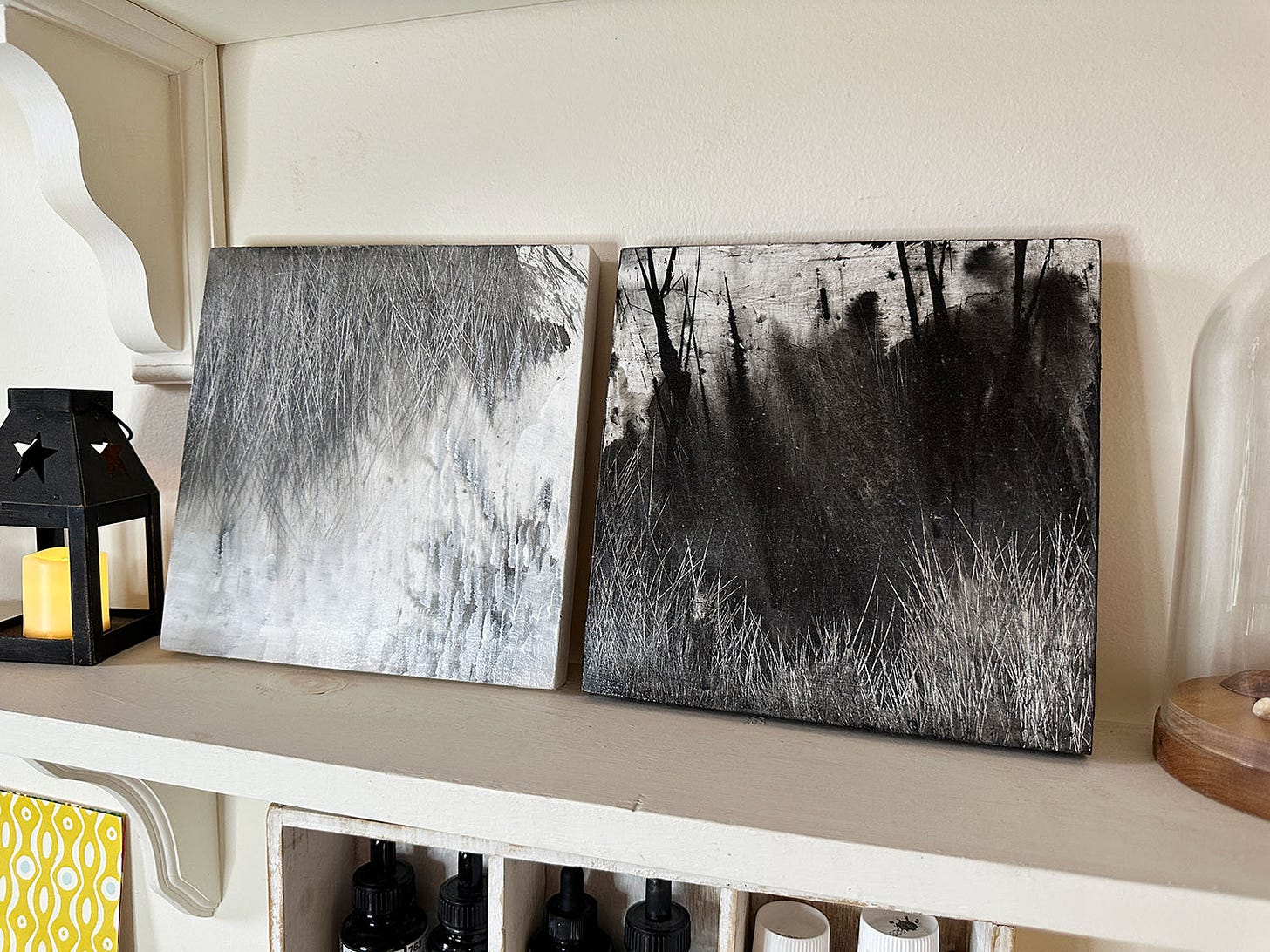 A pair of small wooden panels, one pained in white and light grey, the other primarily in black; they capture abstracted landscape imagery of grasses and branches. Paintings by E Daggar.