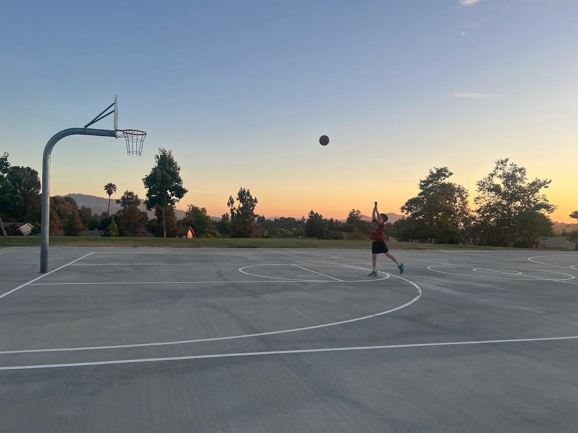 Photo of a boy shooting a basketball on an outdoor court at sunset with trees and mountains in the background.