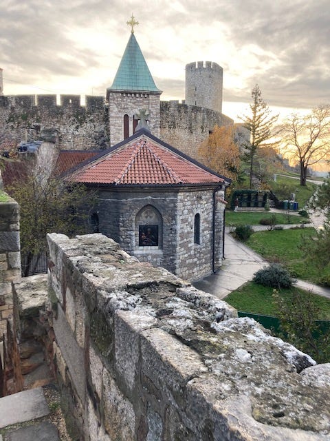 View of medieval fortress: a stone wall, an octagonal chapel, a church spire, a tower. Beyond are sunlit clouds.