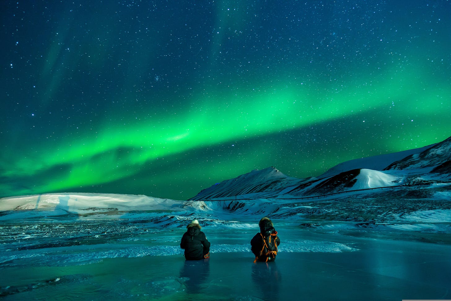 Two figures in warms clothing standing waist-deep in a lake with snowy slopes in front of them. The sky is swirling with the blues and greens of the aurora.