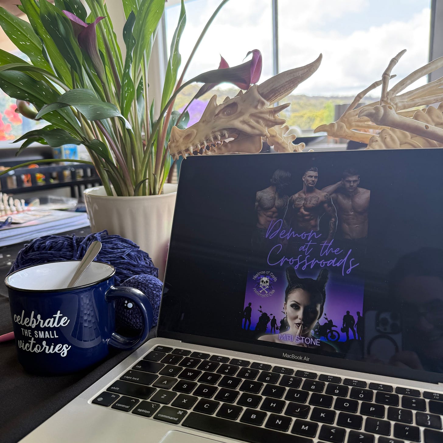 Candid new release photo of Miri Stone's 'Demon at the Crossroads' where the book cover is showing on laptop screen next to a pot plant and a mug which reads 'Celebrate the small victories'