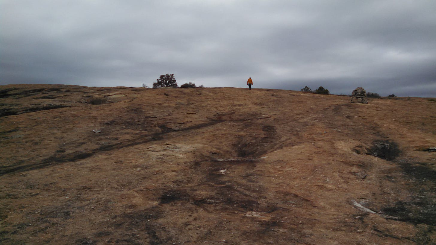 A small figure in a yellow jacket stands on top of a slope of mostly smooth, flat stone, under a cloudy, gray sky