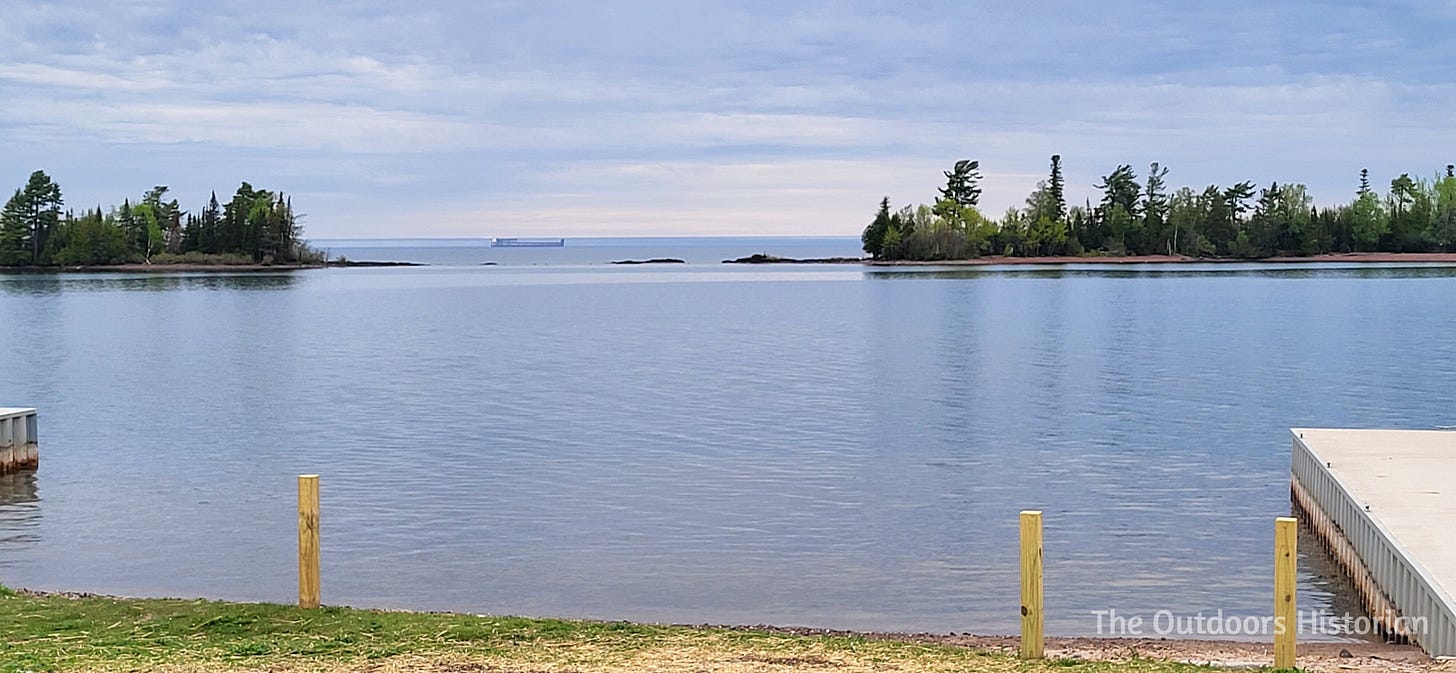 A freighter is seen in the distance passing by the opening of the two points that form Copper Harbor. The lake is calm and the sky is cloudy.
