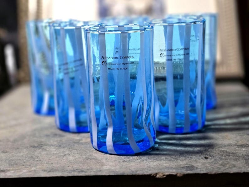 May include: A set of clear blue drinking glasses with vertical white stripes. The glasses have a cylindrical shape and are made of glass. The text "Alessandro Coppola" and "Made in Italy" is visible on the side of the glasses.