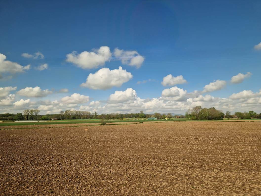 Picture out of the train window. Flat, a brown field, then some green grass and trees; then nice individual clouds and blue skies. 