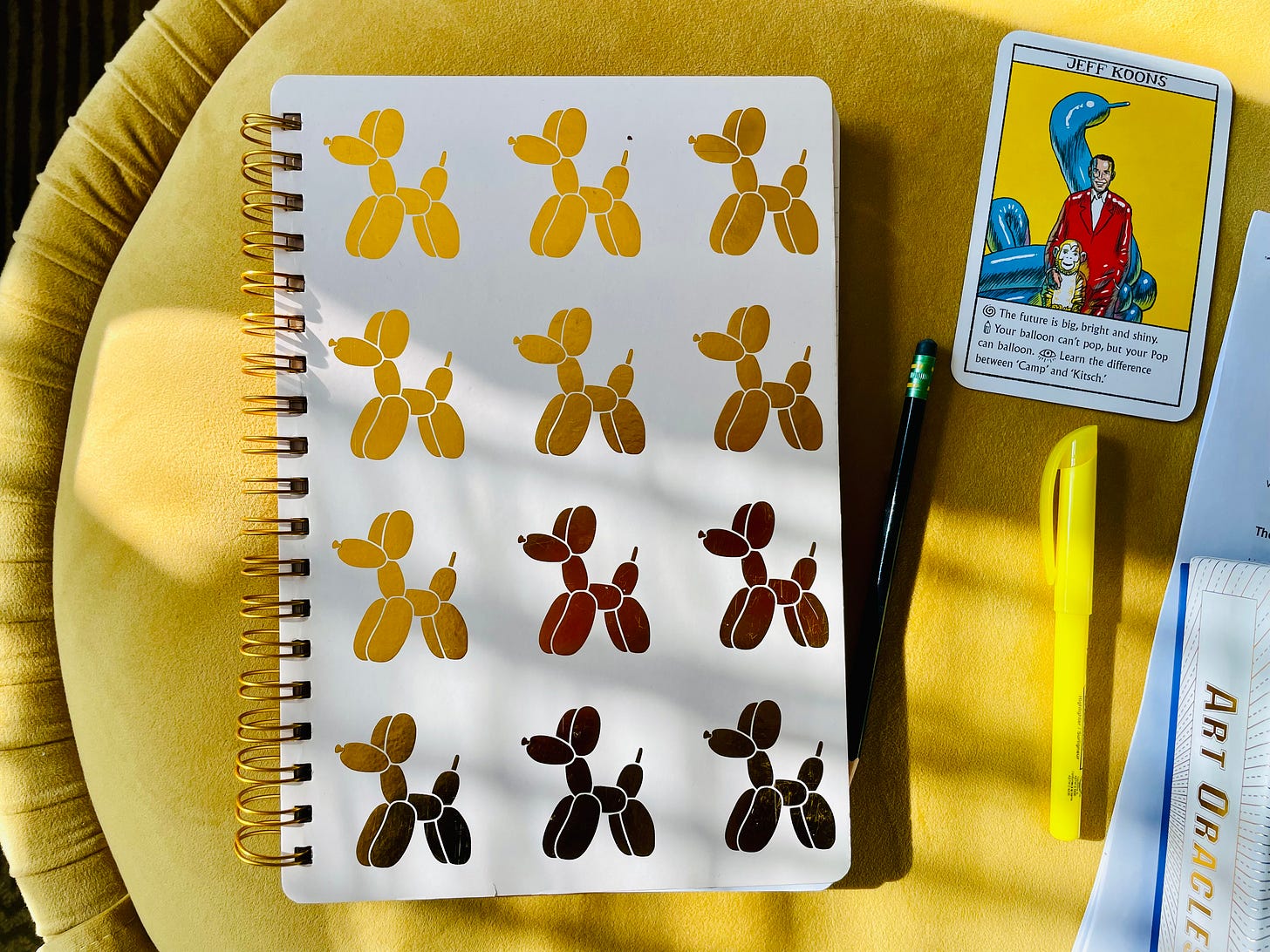 The surface of a yellow ottoman, and on it, a white notebook with twelve gold-foiled silhouettes of Koons' balloon dog. There is a pencil and yellow highlighter beside the notebook.