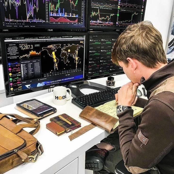 This may contain: a man sitting at a desk in front of two computer monitors with multiple screens on the wall