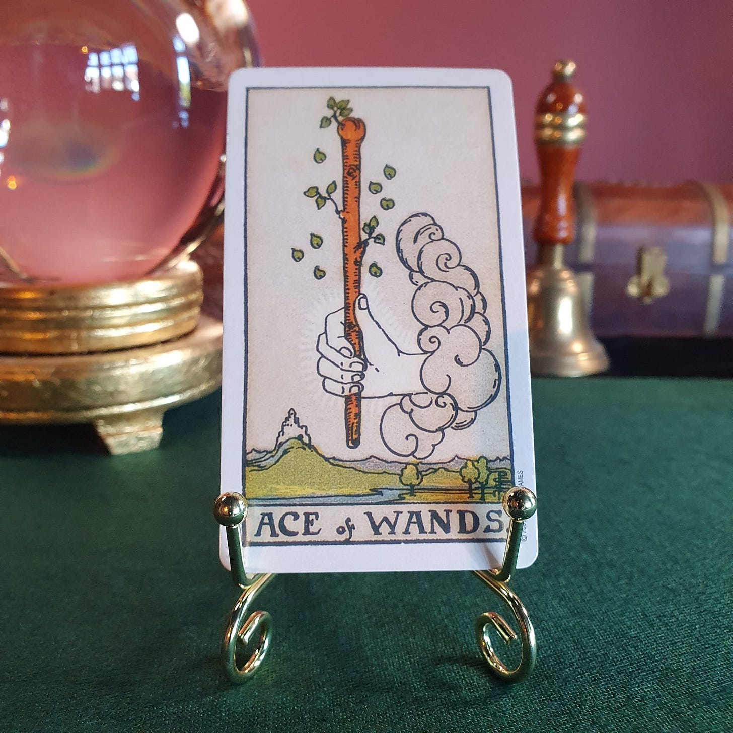 The Ace of Wands tarot card from the Smith-Waite deck displayed on a small gold stand, with a crystal ball, bell, and wooden chest in the background.