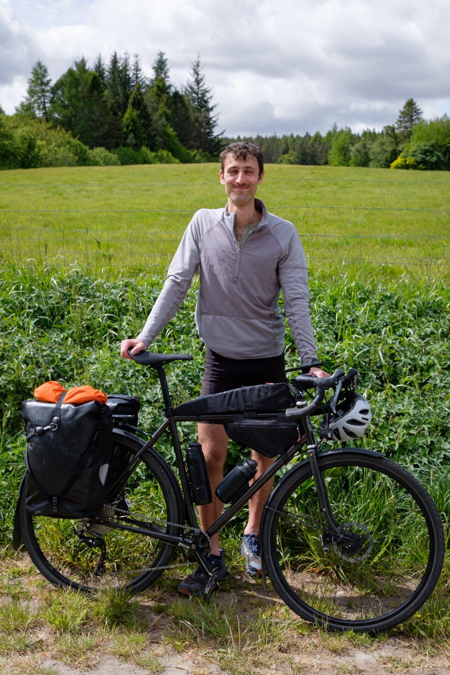 Standing beside a flowing field in the Scottish countryside, I hold my touring bike and smile softly. The landscape stretches out behind me, full of green and golden tones, as I return home with a quiet sense of belonging.