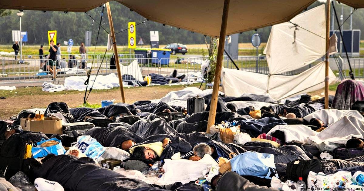 People sleeping on the ground in a temporary shelter, surrounded by personal belongings and a makeshift tent, highlighting the challenges faced by displaced individuals.