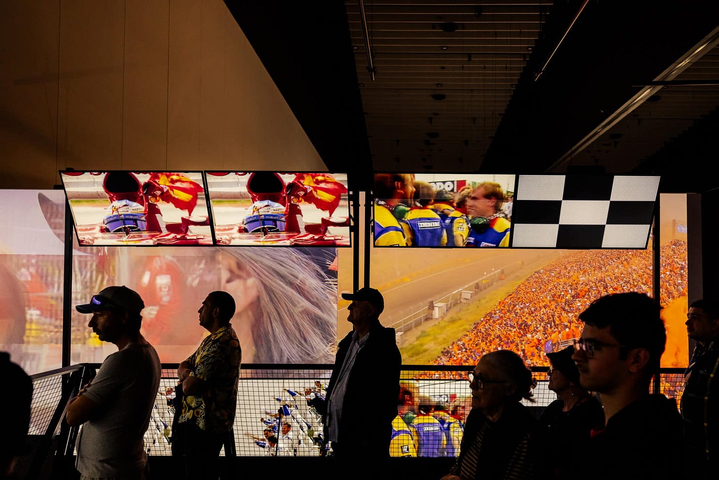 Visitors stand in silhouette inside a dark exhibition space, facing a large wall of video screens displaying colorful motorsport footage, including race cars, pit crews, a packed grandstand, and a black-and-white checkered flag. The bright displays contrast with the dim room, creating a dramatic, immersive atmosphere.