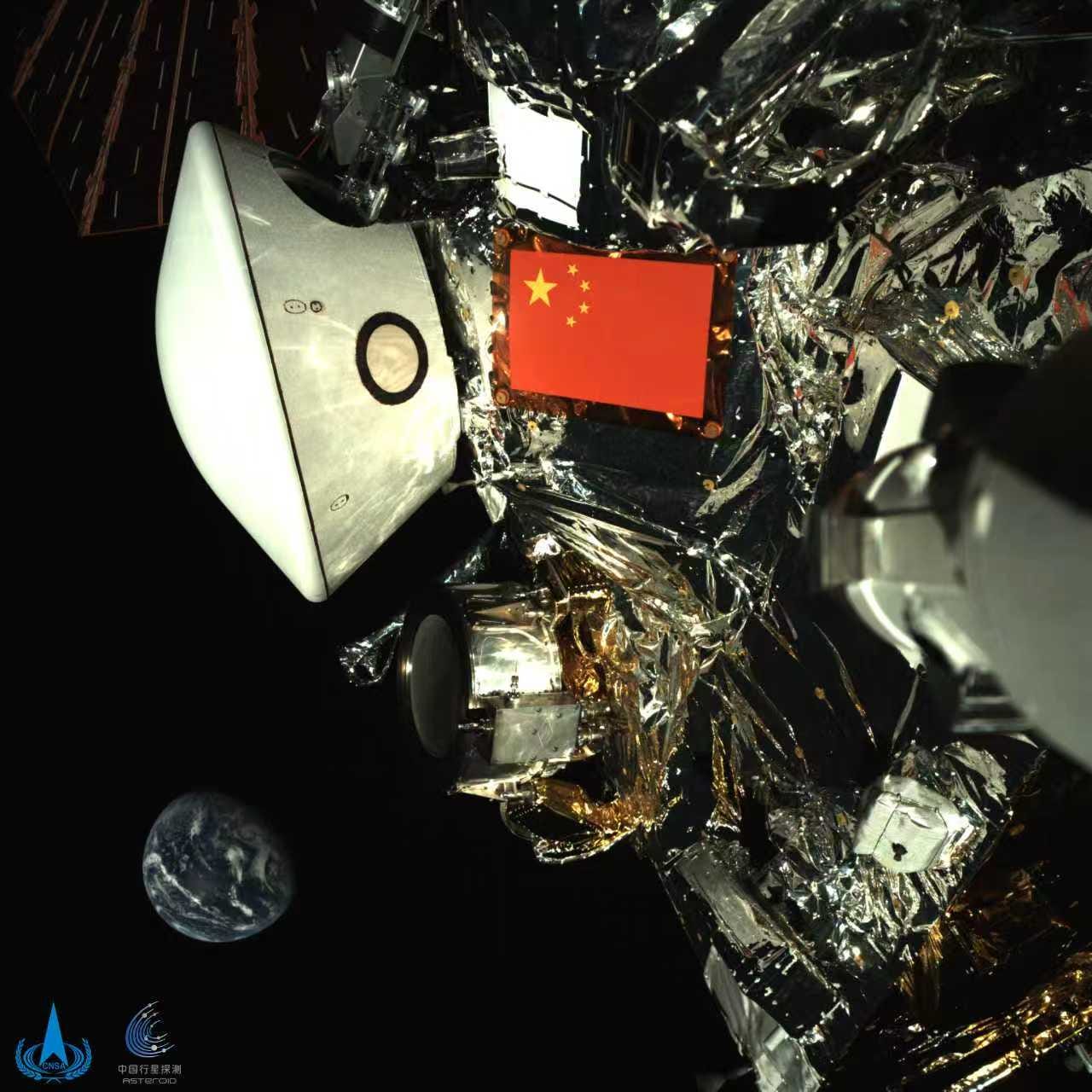 An image from Tianwen-2, released October 1st, shows its sample return atmospheric entry shell next to a flag of the People’s Republic of China, with Earth in the lower left. An image from Tianwen-2, released October 1st, shows its sample return atmospheric entry shell next to a flag of the People’s Republic of China, with Earth in the lower left.