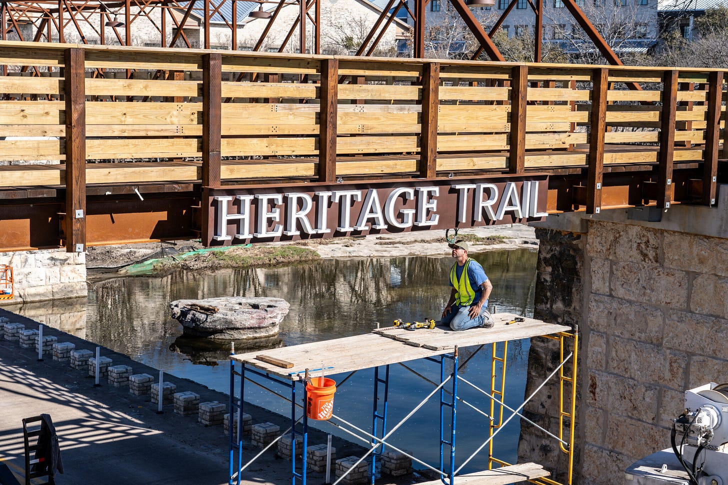 Construction worker kneels on scaffolding while installing a large “Heritage Trail” sign beneath a new pedestrian bridge near downtown Round Rock, with a creek and limestone outcroppings below.