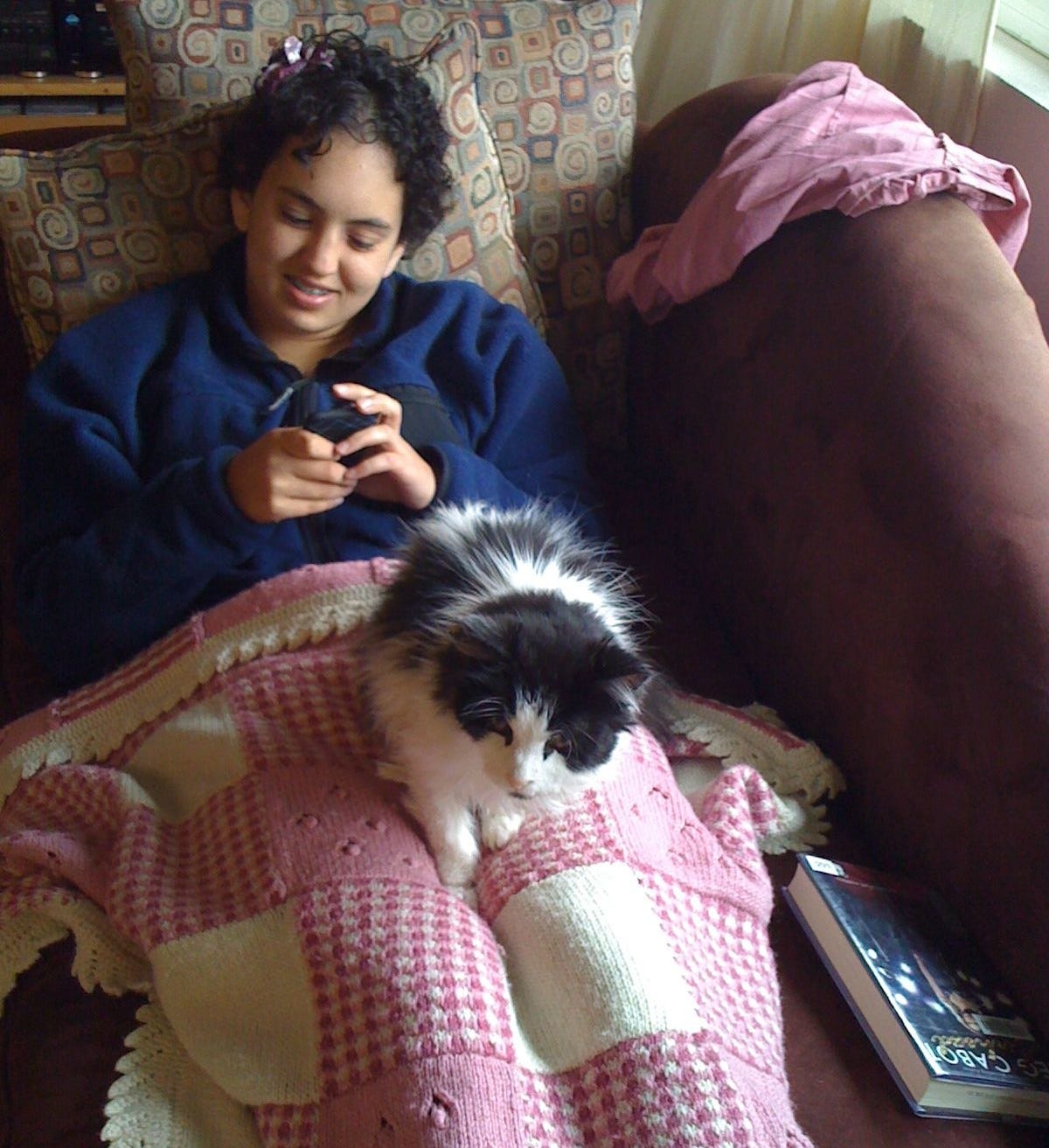 Spot rests his black and white floof contentedly on a blanket, which is spread over a lap on a couch. Both cat and person are cozy and at ease, sharing a quiet, happy moment together. Spot rests his black and white floof contentedly on a blanket, which is spread over a lap on a couch. Both cat and person are cozy and at ease, sharing a quiet, happy moment together.