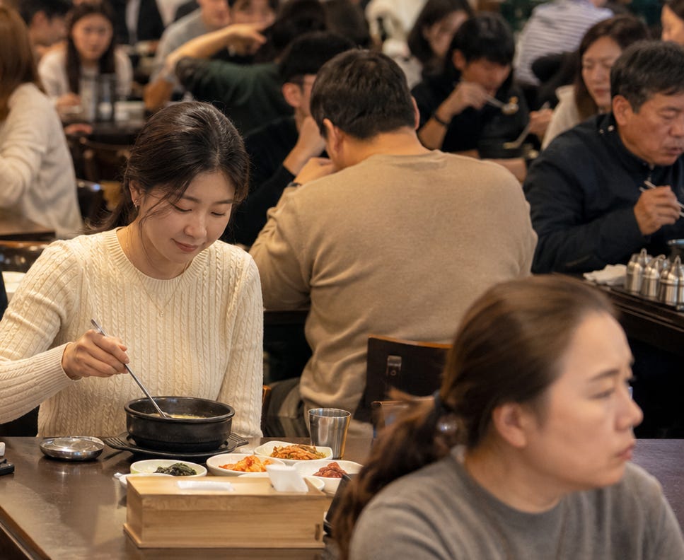 A photo-realistic image of a crowded Korean restaurant filled with diners in conversation. At the center, a woman in a cream sweater eats alone at a small table with a bowl of soup and several side dishes. She appears calm and comfortable, showing solo dining as an ordinary, natural part of everyday life rather than a sad or unusual scene. A photo-realistic image of a crowded Korean restaurant filled with diners in conversation. At the center, a woman in a cream sweater eats alone at a small table with a bowl of soup and several side dishes. She appears calm and comfortable, showing solo dining as an ordinary, natural part of everyday life rather than a sad or unusual scene.