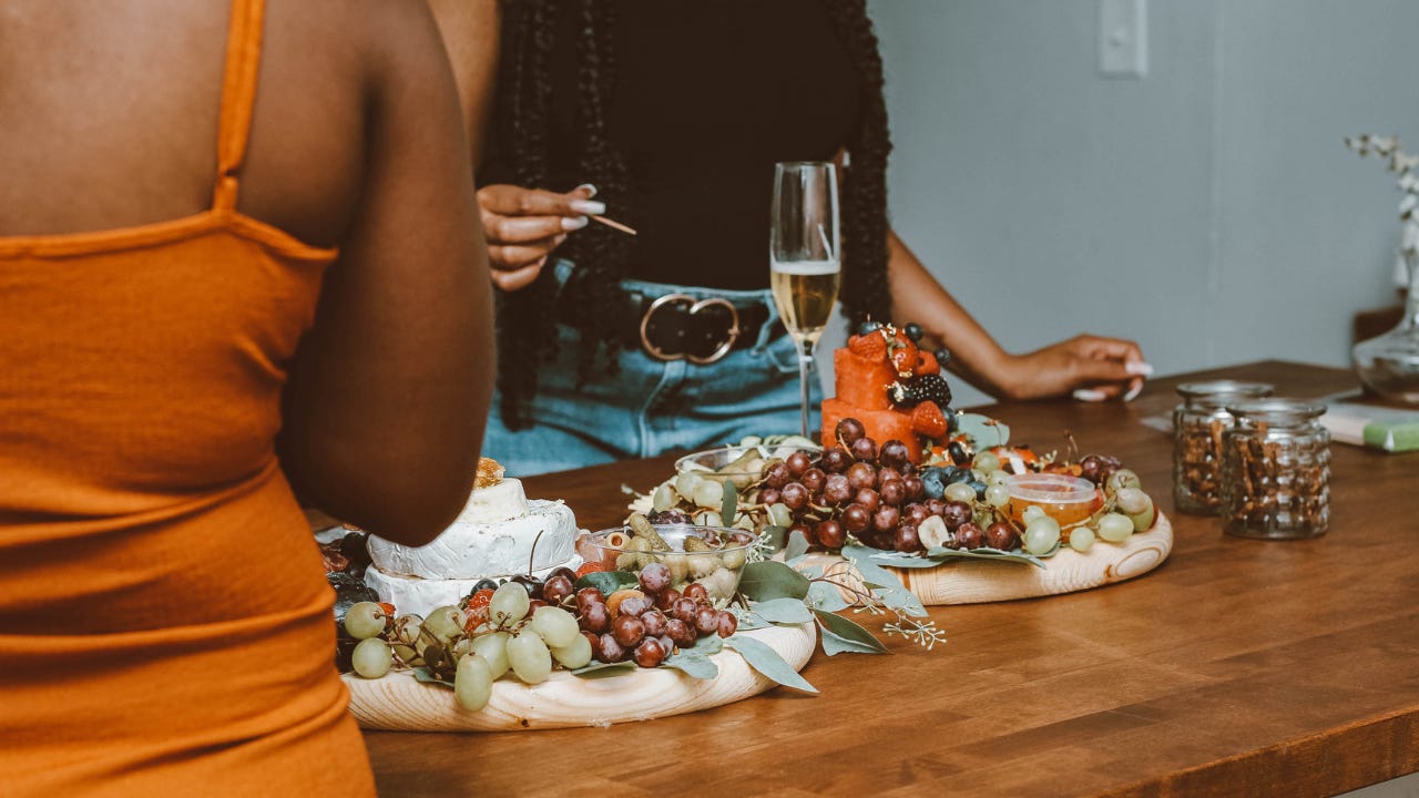 Two Black women stand at a wooden table, joyfully sharing a colorful charcuterie board with cheeses, fruits, grapes, and garnishes.