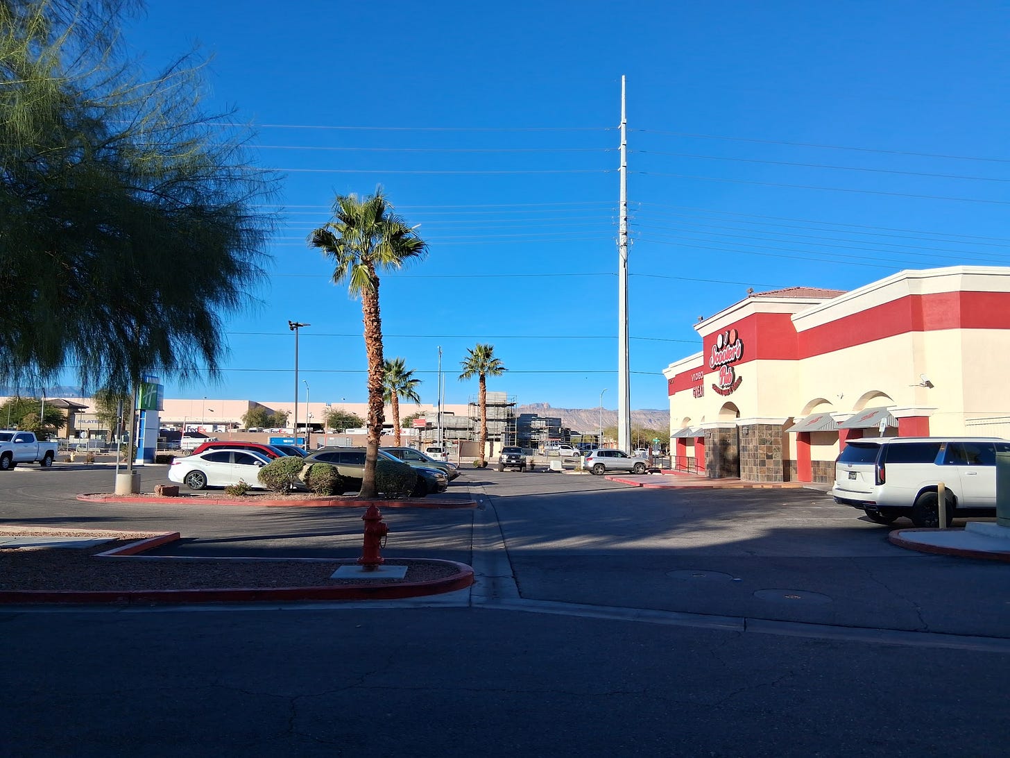 Parking lot with a fast food restaurant on the right side, a palm in the middle, and mountains in the background.