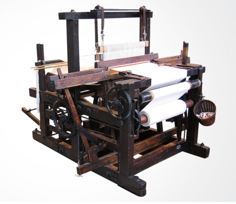 A Toyoda power loom made of dark wood and metal gears, with white fabric partially woven on its rollers and a small basket of spools hanging from the side. The loom’s mechanical parts are exposed, showcasing its intricate, early industrial design.