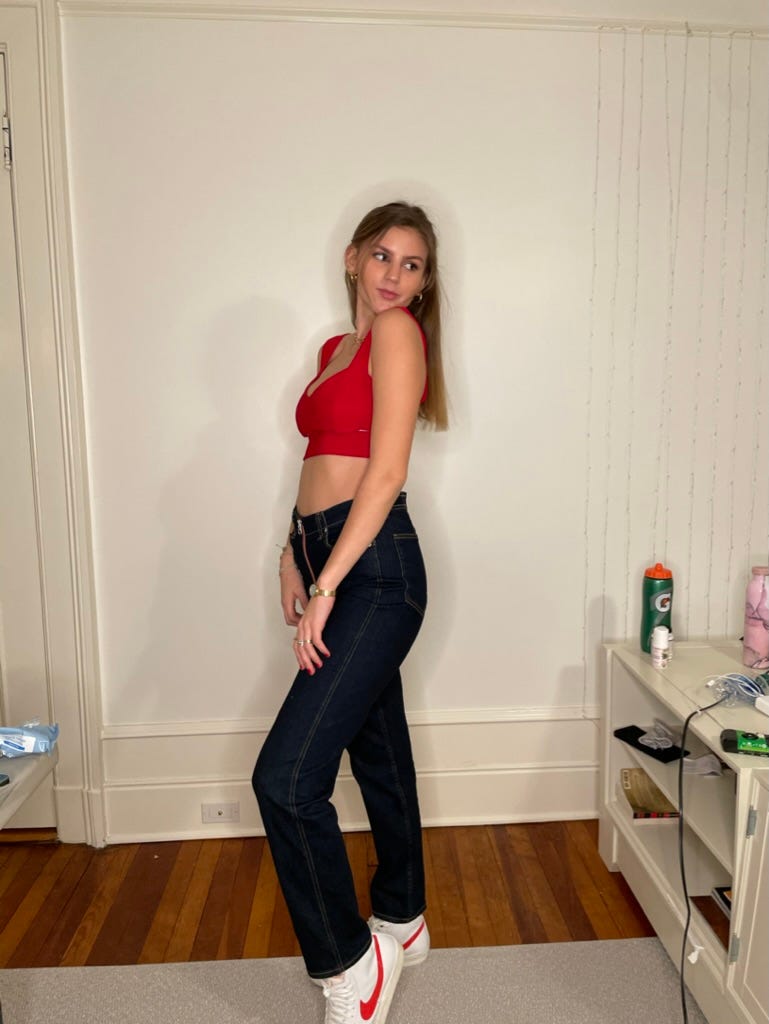 Kat in high school posing for a photo. Their hair is long and they are wearing high rise jeans and a red crop top, looking very femme and also kind of concerned.