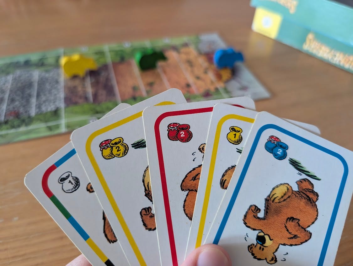 Im Hintergrund ist der Spielplan abgebildet, im Vordergrund sind Karten mit Bären in unterschiedlichen Farben mit einem oder zwei Honigtöpfen. Im Hintergrund ist der Spielplan abgebildet, im Vordergrund sind Karten mit Bären in unterschiedlichen Farben mit einem oder zwei Honigtöpfen.
