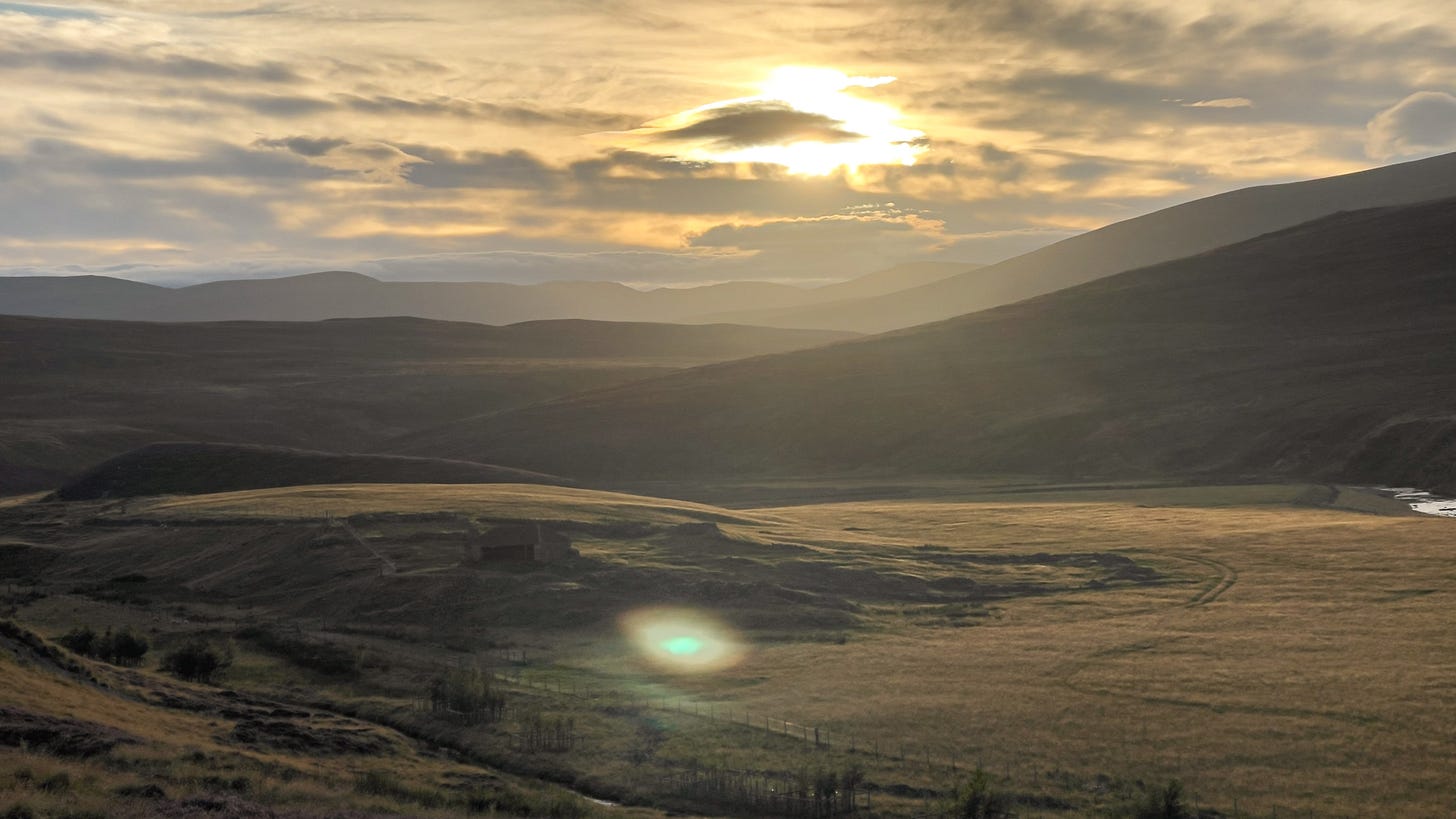A view from high above, looking back across rolling hills layered into the distance. The entire landscape is bathed in golden sunset light, with soft shadows stretching across the land. A peaceful, glowing moment before the final ride downhill.