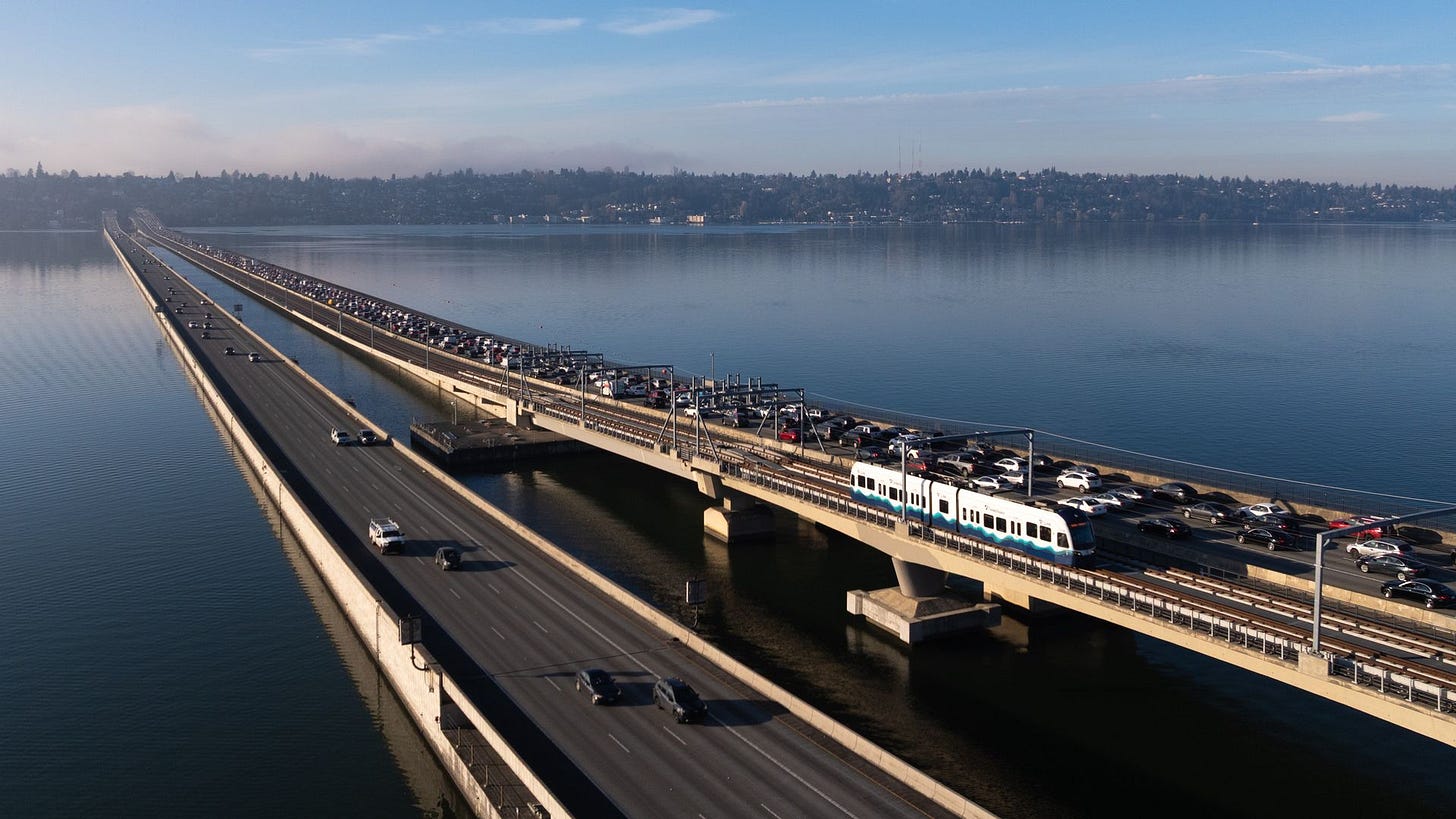 Aerial view of two parallel bridges over a calm body of water. A blue and white passenger train travels on the near bridge while cars line the far roadway, with distant hills under a clear blue sky.