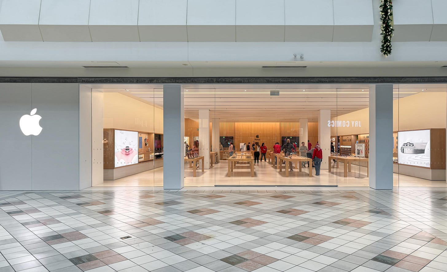 The exterior of Apple South Shore. The store is pictured preparing to open and staff are moving around inside. The mall hallway is brightly lit and empty.