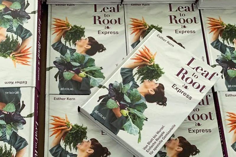 Mehrere Exemplare des Kochbuchs «Leaf to Root Express» von Esther Kern liegen gestapelt in einem Karton. Auf jedem Buchcover ist die Autorin zu sehen, wie sie ein Bündel Karotten mitsamt Grün hochhält und daran riecht, während sie weiteres Gemüse im Arm trägt.