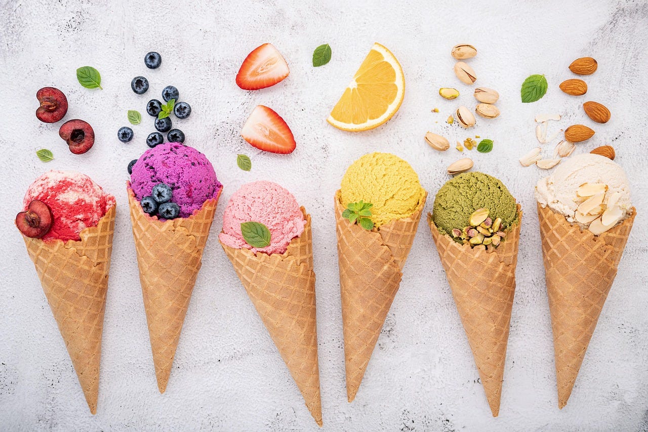 Six ice cream cones of different colors, such as red, purple, pink, and orange, appear in a row with fruits such as cherry, blueberries, and strawberries above them. Some ice cream cones have nuts, such as almonds and pistachios, above them.