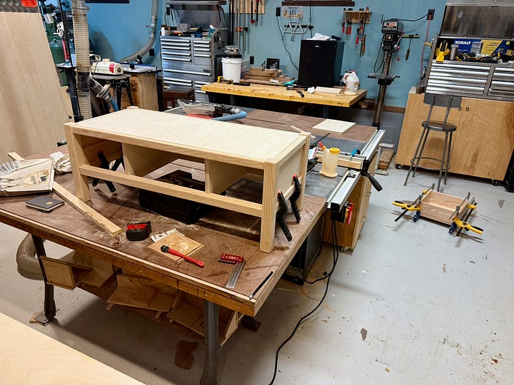 Two workshop views of a wooden bench project. Left: unfinished bench frame clamped together on a workbench. Right: the same bench painted dark, with a drawer box slid partway into place.