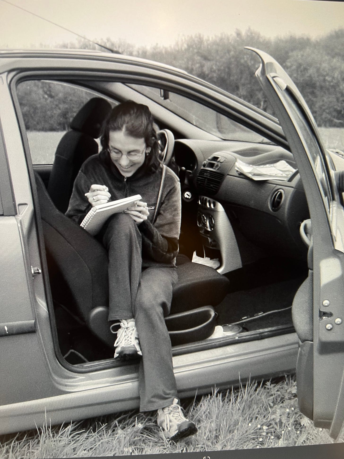 Katy sitting in the passenger seat of a car, door open, writing in a journal. 