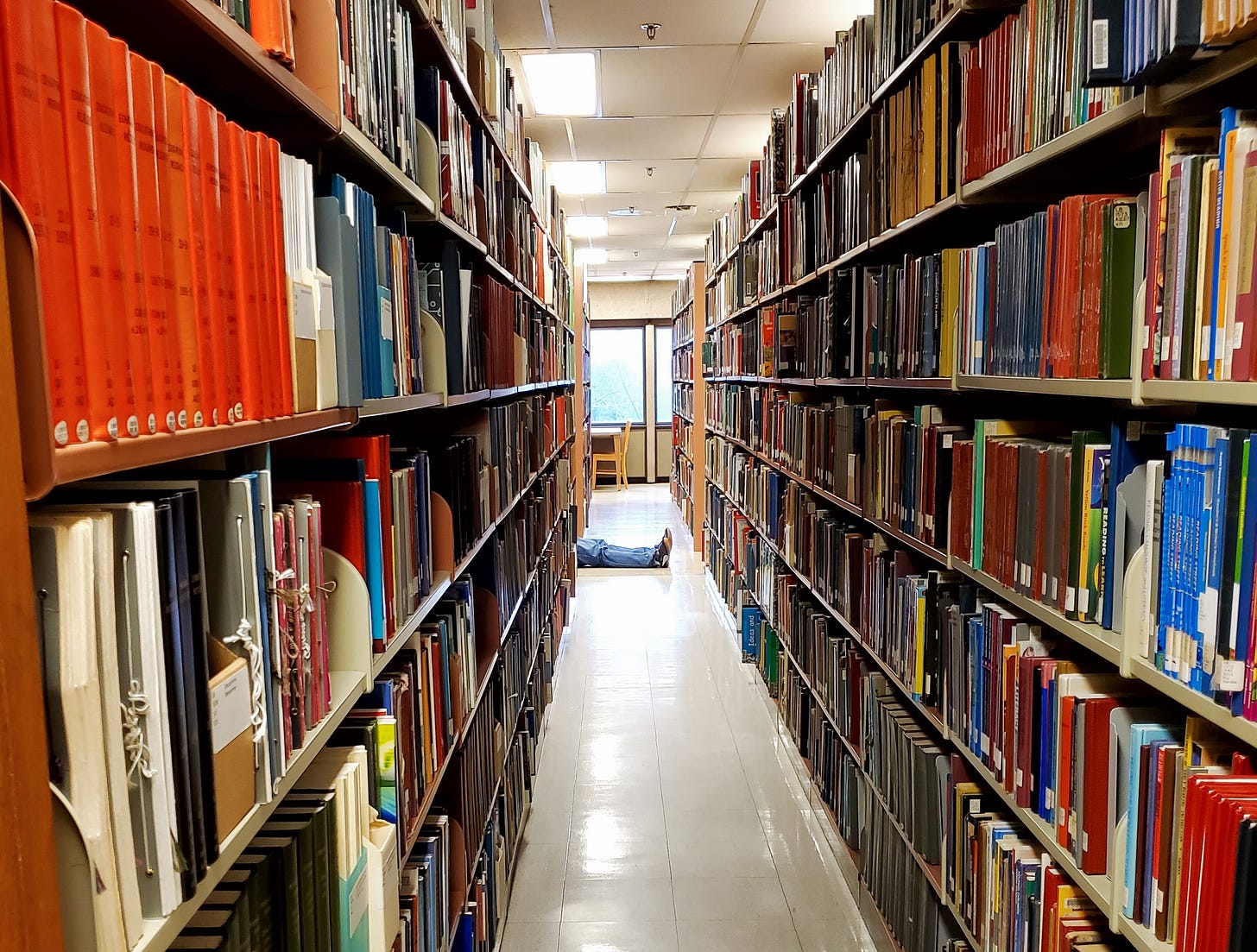 A man’s legs stick out from among the stacks of books at a library.