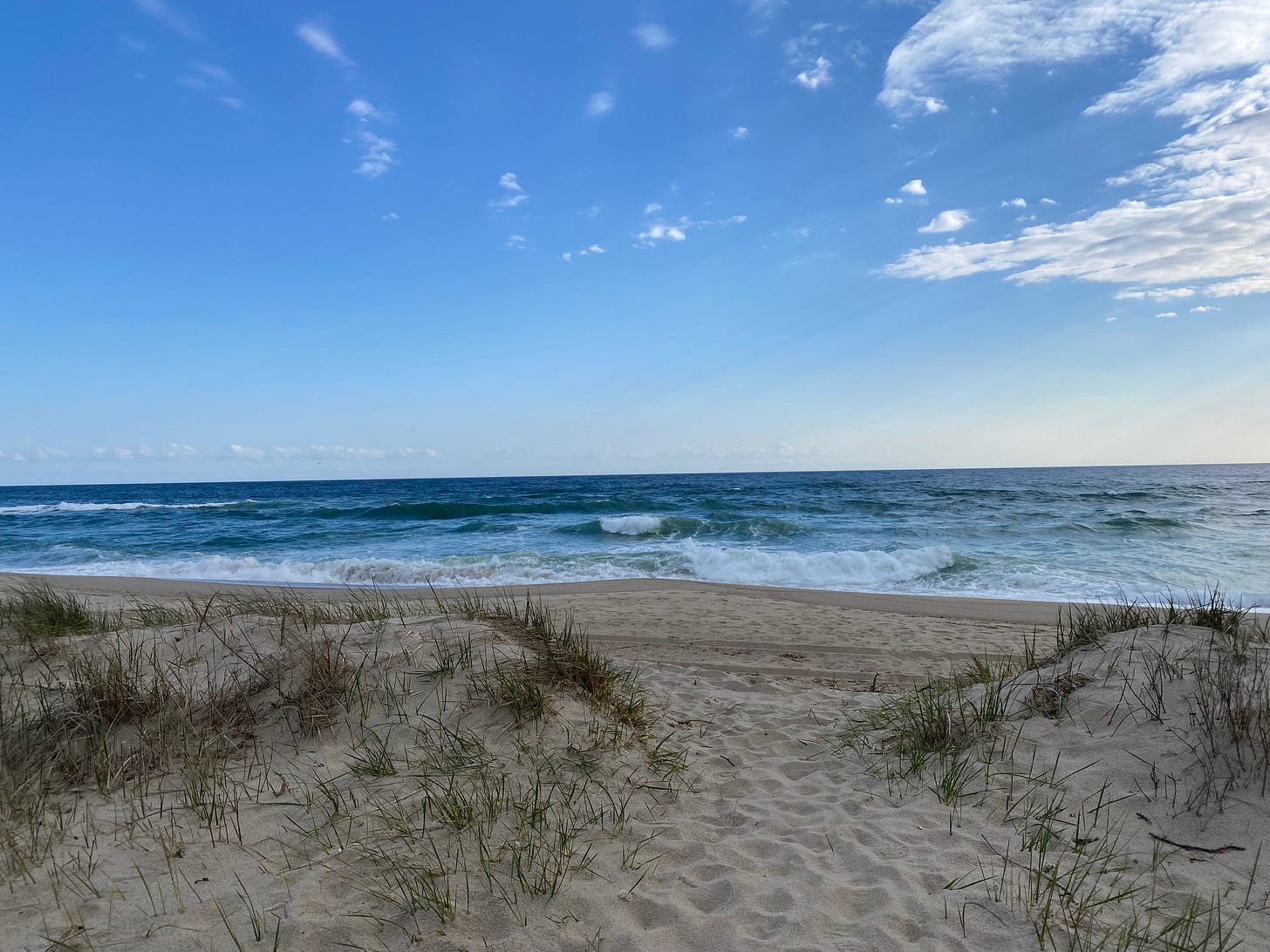 A sandy path through two low dunes, opening onto a wide beach. The ocean is deep blue and full of crashing white waves.